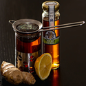 A jar of golden honey, a metal tea infuser resting across a glass of dark amber tea, a half lemon, and a piece of fresh ginger root arranged on a dark tabletop.